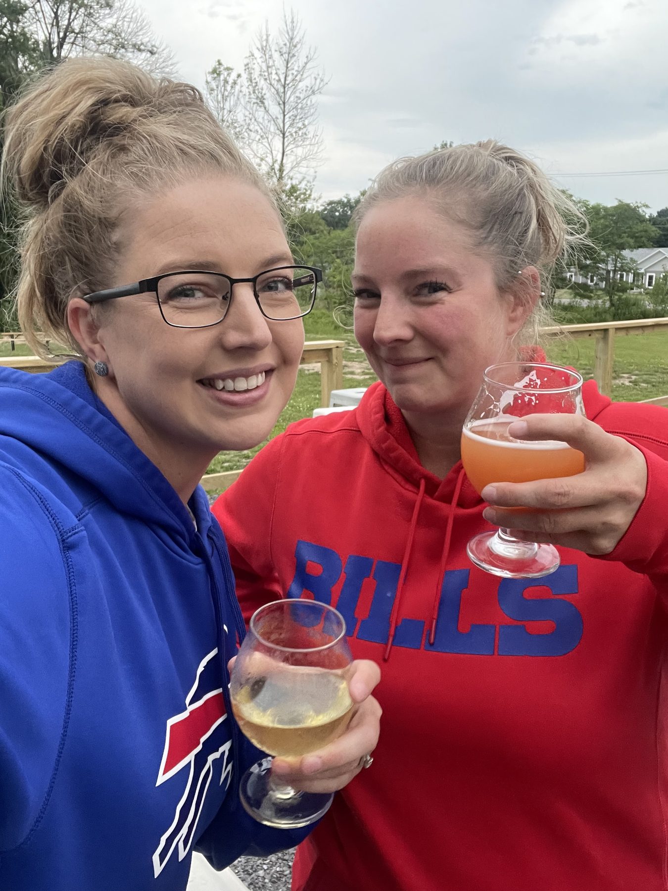 Aleasha and a friend in Buffalo Bills hoodies raising glasses at an outdoor table.